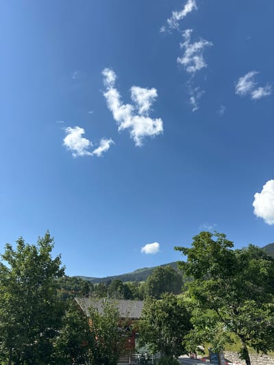 The image depicts a scenic view in Hasliberg Reuti, Switzerland, characterized by a clear blue sky dotted with fluffy white clouds. In the foreground, lush green trees dominate the scene, with their leaves rustling gently. Behind the trees, a rustic wooden building with a tiled roof is visible, suggesting a dwelling or a local establishment. Further in the background, rolling hills covered in dense forest extend towards the horizon, creating a picturesque mountain landscape. The overall atmosphere is tranquil and idyllic, capturing the natural beauty of the Swiss Alps.