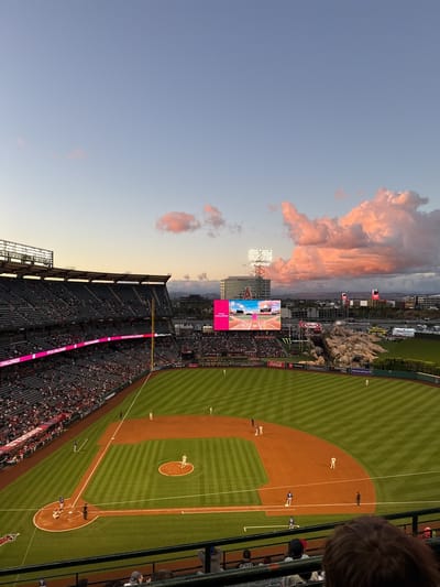 This wide-angle image captures a baseball game in progress at Angel Stadium in Anaheim, California, during the twilight hours. The sky transitions from a clear, pale blue at the top to warm orange and pink tones near the horizon, illuminated by a sunset that casts a vibrant glow on several fluffy pink clouds.

The main subjects are the baseball field and the surrounding stadium. The manicured green outfield features distinct striped patterns, while the brown dirt infield is clearly delineated by white bases and foul lines. Several baseball players are visible on the field in both light and dark uniforms, positioned for play; a pitcher is on the mound, a catcher is behind home plate, and other fielders are strategically placed.

The stadium stands are packed with spectators, particularly on the left side of the frame, indicating a well-attended game. A large video scoreboard in the background displays imagery related to the game or advertisements. To the right of the scoreboard, a distinctive rock formation with artificial waterfalls, a signature feature of Angel Stadium, is visible. Tall stadium light towers are also seen, beginning to illuminate as natural light fades.

Notable details include various sponsor logos and text. A ribbon board prominently displays "PITCH CHALLENGED." Other visible brand names include "Modelo," "Coca-Cola," "Monster Energy," "Yakult," "Toyota," "Yokohama Tires," and "Coors Light." A red "A" logo, likely representing the Angels team, is visible on a building in the distance. The bottom right foreground shows the top of a spectator's head, indicating the viewpoint from the stands. The scene conveys the excitement of an evening baseball game under a colorful sky.