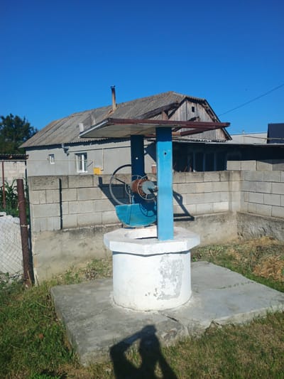 This image, taken in the city of Mitoc, Moldova, depicts a well in a rural or semi-rural setting. The well has a white, concrete base and a blue structure supporting a mechanism for drawing water, which includes a wheel and a rope or chain. Behind the well, a grey, cinder block wall separates it from a building with a weathered, grey, corrugated roof and visible wooden beams. A chimney extends from the roof of this building. The area around the well is covered with grass and dirt. The sky above is clear and blue. A fence made of chain-link and metal posts is visible to the left of the well. The shadow of a person holding a camera is visible in the foreground, indicating the photographer's presence.