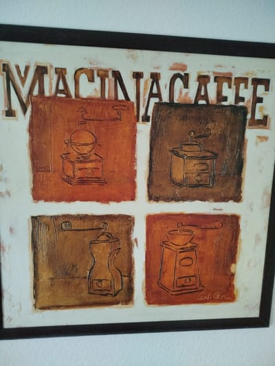 This is a framed painting of a coffee grinder with the text "MACINACAFE" at the top. The painting is divided into four quadrants, each depicting a different coffee grinder. The colors used are shades of brown and orange, with a textured background. A signature is visible at the bottom right corner, appearing to read "Conle Cern". The painting is hanging on a white wall, with a dark frame. There is no discernible connection to Kitzingen, Germany.