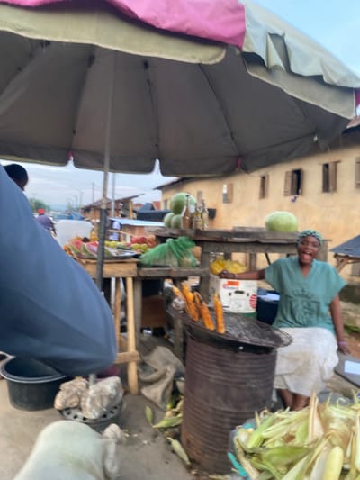 This image depicts a bustling street market scene in Igarra, Nigeria, viewed from a slightly obscured, close-up perspective. In the center-right, a dark-skinned woman, identified as a street vendor, is seated behind her stall. She wears a green V-neck shirt, a white skirt or wrapper, and a patterned green and black headscarf. Her expression is one of broad mirth, with her mouth open in a laugh, and she is looking directly towards the camera. Her right arm is extended, holding a yellow cob of corn over a charcoal grill, which consists of a round metal grate placed atop a rusty metal drum. Several cobs of roasted corn are already on the grill.

The vendor's wooden stall is laden with various produce. Visible items include multiple large green watermelons, smaller fruits (possibly oranges or apples), and produce contained within green plastic bags. Several bottles, possibly containing drinks or oil, are also on the stall. A white box with a red and white logo is visible beneath the stall. On the ground to the right of the grill, a pile of discarded corn husks is present.

Overhead, a large market umbrella, colored in sections of pink and light khaki/green, provides shade, its grey underside visible. The upper left foreground is partially obscured by the dark blue-clad shoulder and arm of an unseen individual, beneath which a black bucket and a light-colored object are on the ground.

In the background, behind the vendor's stall, stands an old, light brown-colored building with multiple rectangular windows, some featuring open or closed wooden shutters. Beyond this structure, a street with indistinct vehicles and other simple market structures or buildings can be seen under an overcast sky, suggesting an outdoor market setting within the city.