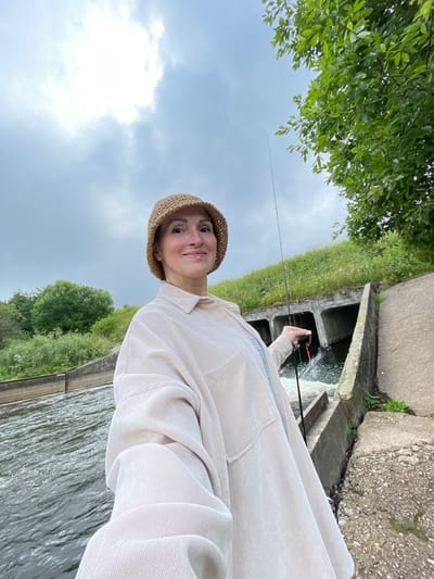 In the city of Bereznik, Russia, a woman stands on a concrete embankment next to a body of water. She is wearing a tan bucket hat and a light-colored, oversized shirt. She holds a fishing rod in her right hand, and a fishing lure is visible on the line. Behind her, a concrete structure with multiple openings allows water to flow through, creating a small waterfall. The surrounding area is covered in green grass and trees, under a cloudy sky. The overall scene suggests a person engaged in fishing by a water channel.