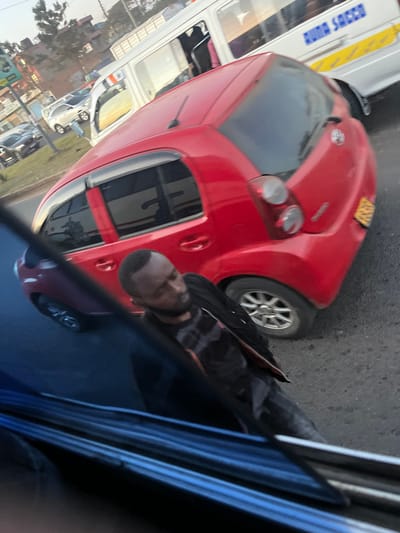 The scene depicts an urban street in Biashara ward, Kenya, under natural daylight conditions. A red five-door hatchback automobile, possibly a Toyota Passo model, occupies the mid-ground, positioned at an angle with its rear and passenger side visible. The vehicle features tinted rear windows and a yellow rectangular license plate affixed to its rear bumper. To the immediate left of the red car, a male individual with a short beard is observed walking on the asphalt surface. He is wearing a dark, possibly black, zipped jacket over a dark-patterned shirt and dark trousers. His gaze is directed towards his left. In the background, a white commercial van or minibus, identifiable by "BUNA SACCO" text and yellow striped decals on its side, is visible. Further in the background, other vehicles, including a white SUV and a dark sedan, are present on a multi-lane road alongside a grassy median and distant urban structures under a clear sky. The image is captured from the interior of a vehicle, with a dark blue portion of its frame and reflections of the street scene visible in the foreground.