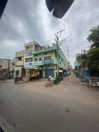The image captures a street scene in Bethamcherla, India, under a cloudy sky. The perspective suggests the photo was taken from a vehicle, looking out at a busy urban environment. The street is paved and appears worn. On either side of the street are multi-story buildings, primarily shops and residences.
The buildings display a range of architectural styles and colors, with some painted in vibrant greens and others in more muted tones. Many of the ground floors house commercial establishments with visible signage in what appears to be Telugu script, indicating shops such as "BSR FASHION HUB," "MY DR," and a general store or bazaar. Awnings extend over some shop fronts.
Several individuals are present in the scene. A man is seated on a motorbike in the foreground, while others can be seen walking or standing near the shops. One person is seated on a bench outside a shop to the right. There are also various objects indicative of street life: a flower stall with colorful garlands, bicycles, and what appears to be a handcart. Electrical wires and poles are prominent overhead, typical of many Indian urban settings. In the distance, a telecommunications tower is visible. The overall impression is one of a functioning commercial street in a moderately populated town.