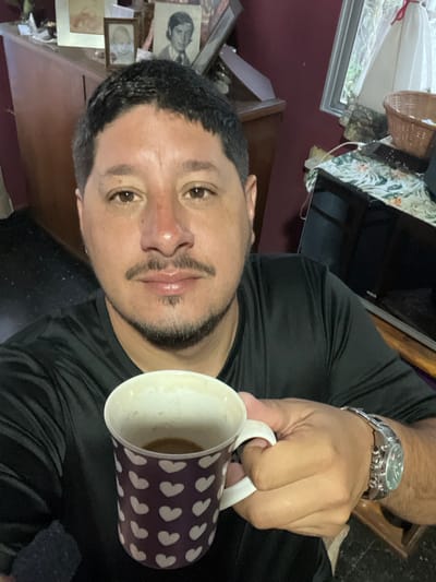 An adult male is centered in a close-up, chest-up selfie taken indoors in Bernal Este, Argentina. He has short dark hair, a mustache, and a short beard. He is wearing a black short-sleeved t-shirt and a metallic wristwatch on his left wrist. His right hand holds a dark purple ceramic mug with white heart patterns, containing a small amount of light brown liquid. The man looks directly at the camera with a neutral expression. In the background, to his left, is a wooden cabinet displaying multiple framed photographs, including one of a baby and one of a young man. To his right, a window with a white curtain is visible, along with a woven basket and a black-screened television set resting on a surface covered by a floral-patterned cloth. The room's walls are painted a dark red.