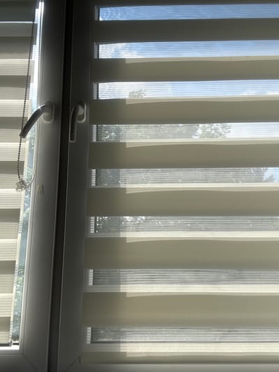 The image shows a close-up view of a window with dual-layer blinds, a common type of shade that alternates between opaque and sheer fabric. The blinds are partially open, allowing light to filter through. The window frame is white, and on the left side of the frame, a beaded cord mechanism is visible, used for operating the blinds. A white window handle is also present on the left side.
The outdoor environment visible through the blinds suggests a daytime scene with a blue sky and some clouds. Trees are also visible, indicating an outdoor setting. The lighting suggests it is daytime with sunlight. The image composition focuses on the details of the window and blinds, with the outdoor environment serving as a backdrop.
The provided information states this image is located in the city of Kalisz, Poland. Without further context or visual cues within the image itself, it is impossible to definitively confirm this location. The scene depicts an interior view of a window, which could be found in any residential or commercial building.