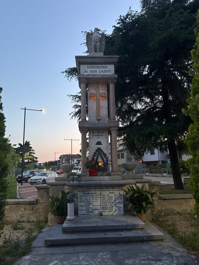 The image shows a war memorial in Dossobuono, a frazione of the city of Villafranca di Verona, Italy, dedicated to those who died during World War II. The memorial consists of a stone structure with pillars, inscriptions, and a statue of an eagle on top. The inscription "DOSSOBUONO AI SUOI CADUTI" (Dossobuono to its fallen) is visible, along with "IN LUMINE GLORIAE" (In the light of glory). A plaque lists the names of those who went missing in the sea and in Russia, during the second World War. The background reveals a street with parked cars and buildings. The sky is visible at dusk. Trees surround the memorial. A street light is visible.