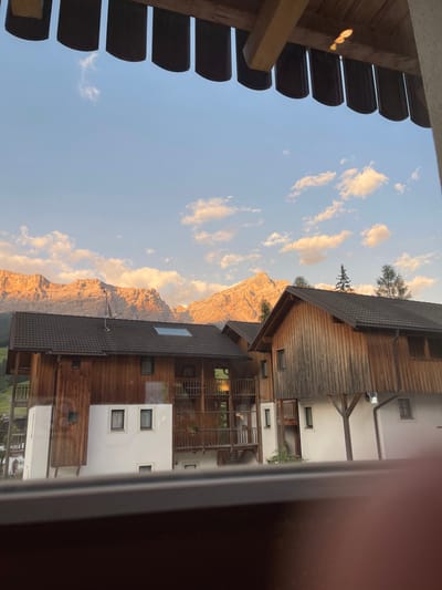 This media file captures a serene mountain landscape in La Ila - Stern - La Villa, Italy, likely during the late afternoon or early evening. The setting features traditional alpine architecture with wooden and white-washed buildings, characterized by multiple levels, balconies, and small windows. One building prominently displays a dark, tiled roof and wooden exterior walls, while the adjacent structure has a white facade with decorative window frames.
The dominant natural subject is a majestic mountain range, its rocky peaks illuminated by the warm, golden light of the setting sun. The mountains are etched with dramatic shadows, highlighting their rugged texture. Scattered clouds dot the clear blue sky, tinged with the soft hues of sunset.
In the foreground, a portion of a wooden structure with a slatted overhang is visible, suggesting a view from a balcony, porch, or window. The composition is oriented vertically, with the sky and mountains occupying the left side and the buildings dominating the right.
There are no people visible in the frame. The scene evokes a sense of tranquility and natural beauty, typical of a mountain retreat. The time of day is suggested by the warm, low-angled sunlight casting long shadows and illuminating the mountains with a golden glow. The weather appears clear and pleasant. No text is visible in the image.