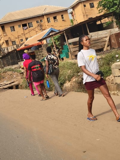 The scene depicts a group of five individuals, appearing to be young adult females, walking along a dirt path bordering an asphalt road in Puerto López, Colombia. In the foreground, a woman of apparent African descent is walking towards the right, her body angled slightly forward, and her head turned towards the left, looking directly into the general direction of the camera. She wears a white short-sleeved t-shirt with a yellow graphic, a dark red mini-skirt, and blue and white flip-flops. A small black shoulder bag hangs on her left side. Her hair is dark and braided.
Behind her, four other individuals are walking away from the camera. The first of these wears a dark, patterned blouse, dark trousers, and dark sandals, carrying a blue water bottle in her right hand and a dark bag over her shoulder. Following her, another individual wears a black t-shirt with a red, yellow, and green graphic on the back, pink shorts, and dark flip-flops. Behind this person, another wears a bright red t-shirt, pink trousers, and a purple head wrap. The fifth person is largely obscured by the individual in red.
The environment includes a prominent light brown, multi-story building with a lighter brown roof situated on the left side, featuring multiple windows, some with security bars, and visible laundry hanging. In front of this building, a smaller, less structured dwelling with a blue roof is present. Overgrown vegetation, wooden structures, and debris are visible along the dirt path and behind the walking individuals. The ground consists of both packed dirt with visible tire tracks and a segment of asphalt road in the lower left. The sky overhead appears hazy.