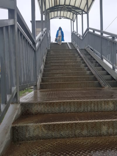 A person wearing a blue hooded raincoat and light-colored pants is walking up a metal outdoor staircase in Бұлақты, Kazakhstan. The person is carrying a light-colored bag in their right hand. The staircase is covered by a metal roof supported by pillars, and has metal railings on both sides. The steps are made of diamond plate metal, and appear wet, suggesting it might be raining or has recently rained. The overall environment is overcast and appears to be a pedestrian overpass.