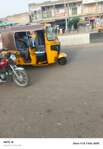 A yellow auto-rickshaw, carrying passengers, moves along a street. A red motorcycle sits to its left. Commercial buildings line the opposite side of the street, with various storefronts visible. The road occupies the majority of the frame, showing wear and some debris. The image is dated "2026.01.27 08:02 AM" and notes camera settings "23mm f/1.8 1/100s ISO93".