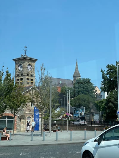 The image depicts a view of the Waterside Train Station in the city of Derry/Londonderry, United Kingdom. A prominent stone clock tower with four clock faces stands tall, topped with a weathervane. To the right of the clock tower, a church spire is visible. In the foreground, a white car is parked, with its front passenger side partially visible. A paved area with benches and trees lines the front of the station. Several people are seen walking and sitting on benches. A blue sign with "North West Transport Hub" is visible. A large billboard displaying an advertisement is positioned behind a black fence. The sky is clear and blue.