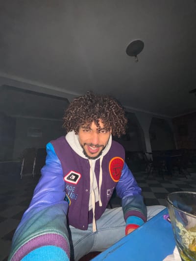 A man with curly hair grins, wearing a purple and blue letterman-style jacket over a white hooded sweatshirt and light-wash jeans. Part of a blue table is visible, with a bucket on top. The background is a dimly lit indoor space with a black and white checkered floor. A light fixture is attached to the ceiling. The man is positioned at an angle, and the image is slightly rotated.