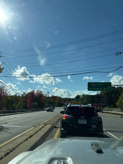 This image depicts a sunny, daytime scene on a multi-lane road in Newton, Massachusetts, United States, specifically on a road approaching I-95 South. The perspective is from the driver's seat of a light-colored vehicle, with its hood visible at the bottom of the frame.
Directly ahead, a black Toyota RAV4, bearing Massachusetts license plate "6RS 491," is stationary or moving slowly in traffic. Additional vehicles are visible in the lanes ahead. The road features multiple lanes separated by yellow markings, and a concrete median strip overgrown with dry vegetation on the left.
Overhead, multiple black power lines traverse a clear blue sky dotted with scattered white cumulus clouds. Bright sunlight from the upper left quadrant causes visible lens flare. On the right side of the road, an overhead green highway sign clearly displays "I-95 SOUTH Providence RI" with a white arrow indicating a rightward turn. A smaller, further sign reads "Exit 17 / Newton St / Wellesley."
The environment includes trees lining both sides of the road, exhibiting autumn foliage with distinct red leaves visible on the left. A utility pole stands on the left side, and distant buildings are visible on the right beyond the highway. No individuals are explicitly visible. The overall scene suggests a congested traffic condition on a major thoroughfare.