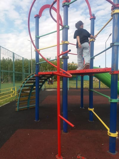 A playground in Бородулиха ауылдық округі, Kazakhstan, features interconnected bars and structures painted in red, blue, green, and yellow. A person in a black shirt and light-colored pants is positioned on the structure, grasping a yellow bar. In the background, there is a green and blue metal fence and sparse trees, suggesting a park-like setting. The ground is covered with a red and black surface, likely rubber matting for safety. In the distance, another playground structure with a person on a swing can be observed. The sky is visible through the playground equipment, showing light clouds.