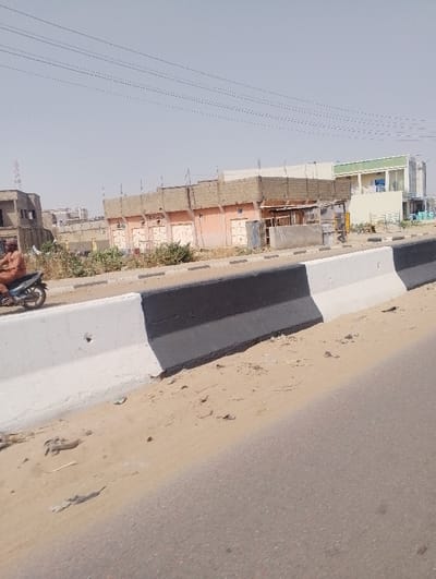 The image depicts an outdoor urban scene in Chiranchi, Nigeria, under a clear, light blue sky. The foreground features a two-lane asphalt road, partially covered by sand on its right side. A concrete median barrier, painted in alternating white and black sections, separates the road from an unpaved shoulder. This shoulder is covered in light brown sand with sparse green vegetation and minor debris.
In the midground, a male individual, dressed in an orange or brown long-sleeved garment, is seated on a dark-colored motorcycle positioned on the sandy shoulder to the left of the barrier. Overhead, multiple thin power lines traverse the frame.
The background consists of various structures. To the left, a multi-story, unfinished building with concrete elements is visible. Adjacent to it, a low-rise structure with multiple light-colored doors or shutters features an upper section constructed from reddish-brown blocks, potentially under construction or in disrepair, with visible framing. Further to the right, a more modern, two-story building with a light yellow ground floor and a light green upper floor, including balconies, stands. Additional indistinct buildings are visible in the far distance, along with a tall, reddish-brown communications tower on the horizon to the left.