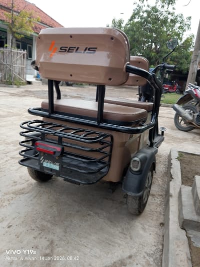 A tan Selis electric bike sits on a concrete surface outdoors. It features a backrest with the Selis logo, a seat for two, and a black metal cargo rack. Behind it, a red motorbike and another motorbike are visible. A building with a red-tiled roof and a bamboo fence are in the background. The lower left corner displays "vivo Y19s, Akhtar & Arvan, 14 Jan 2026, 08.42."