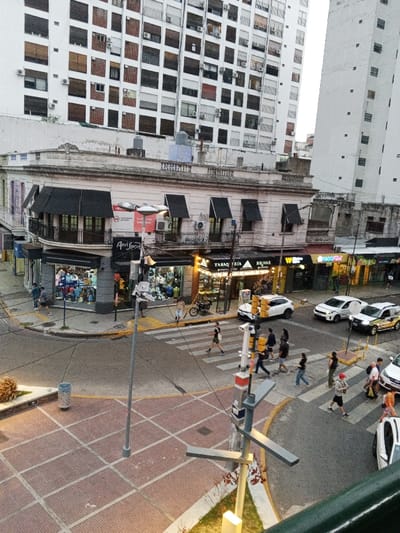 The image presents an elevated view of an urban intersection in Ramos Mejía, Argentina, observed during what appears to be late afternoon or dusk. In the background, multiple white high-rise residential buildings with numerous windows, air conditioning units, and a mix of closed brown and white shutters are prominent.
At the focal point of the midground is a multi-story, light-colored commercial building situated at the street corner. It features an ornate roofline, black awnings over ground-floor storefronts, and balconies on upper levels. Visible shop signs include "Aní Sport," "TABAQUERÍA," "RELMAR," and "PADDY." Adjacent to this building, other commercial establishments line the street.
The asphalt road forms a busy intersection with clearly marked pedestrian crosswalks and yellow curb lines. Sidewalks are paved with square, reddish-brown pavers. A central median area, also paved with reddish-brown tiles and containing some small shrubbery, is visible in the lower left.
Approximately 15-20 pedestrians, dressed in casual clothing, are observed crossing the street at the designated crosswalks or walking on the sidewalks. Several vehicles are present on the road, including a white SUV, another white car, a white taxi-like vehicle with yellow markings, and a partially visible white sedan in the foreground. A bicycle is leaning against a curb near the "TABAQUERÍA."
Street infrastructure includes grey light poles, traffic lights with yellow housings positioned at the intersection, and a grey cylindrical trash bin on the sidewalk. Artificial lights are illuminated within some of the ground-floor shops, consistent with the ambient light conditions.