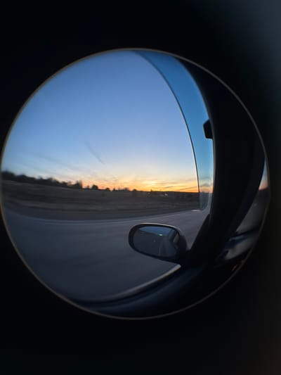 The image shows a distorted view of a sunset landscape, captured through a curved surface that could be a lens or a specialized mirror. The lower half of the image is dominated by a blue sky transitioning into a horizon line marked by golden light, suggesting the sun is setting or rising. The ground is discernible as a flat, grey surface, possibly a road or a field, with darker vertical features, possibly trees or vegetation, separating the sky from the land. The upper part of the image reveals details of a vehicle's interior: the edge of the roof, part of the door frame, and the side-view mirror reflecting the passing landscape. The scene is contained within a circular frame against a black background. The image does not contain any discernible human figures or interactions. The location details of Kuregovo, Russia, are not directly discernible within the image.