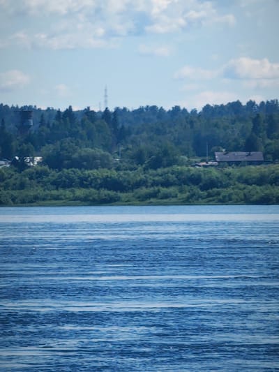 The image shows a wide river with a heavily wooded bank. The river is calm with gentle ripples. The bank is lined with dense green trees and some bushes, creating a lush, natural landscape. In the background, there are a few structures visible through the trees, including a tall tower and a building with a dark roof. The sky is partly cloudy with patches of blue sky visible. The overall scene is tranquil and natural, suggesting a rural or semi-rural setting. It is possible this location is in or near the city of Абалаково, Russia, as indicated.