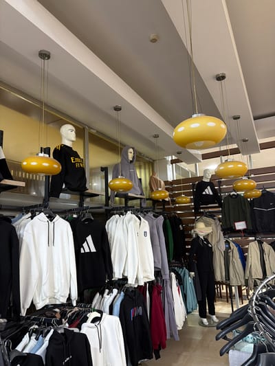The image shows the interior of a clothing store in Acıpayam, Turkey. The store features a variety of clothing, primarily hoodies and jackets, displayed on racks and on mannequins. Several yellow, mushroom-shaped pendant lights hang from the ceiling, illuminating the space. Two male mannequins are visible, one wearing a black hoodie with yellow accents and the inscription "Emirate Fly Biy," and another wearing a grey hoodie. A female mannequin is dressed in a black suit and a straw hat. The store's walls appear to be paneled with wood. Various clothing items are neatly hung on black hangers. The floor is tiled.