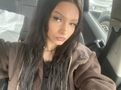 The image depicts a female subject seated inside a left-hand drive vehicle, positioned to capture a selfie. The subject has long, dark hair with lighter highlights, styled to cascade over her shoulders. She wears prominent false eyelashes, winged eyeliner, lipstick, a septum piercing, and two eyebrow piercings on her left brow. Her attire includes a dark inner top and a brown jacket with a distinct fleece texture on the sleeves. A light-colored pearl necklace is visible around her neck.

On the seat immediately behind the subject, a white bag displaying the partial text "OF INNOVATION" is present. Through the vehicle's right-side window, the exterior environment is visible, showing a parked grey vehicle with a French license plate reading "AV-302-W". A dark seatbelt is visible on the right side of the frame. The scene is located in the city of Taverny, France.