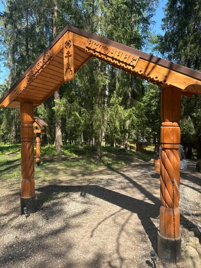 The image shows a wooden archway in a forest setting, possibly in Махра, Russia based on the Cyrillic inscription on the arch. The archway is made of intricately carved wood, with decorative patterns and Cyrillic text. It is supported by two wooden posts with a twisted design. The posts are set into concrete bases. A path leads under the archway and into a forest of tall trees. The ground is covered with gravel and dappled with shadows from the trees. The lighting is bright, suggesting a sunny day. A person is partly visible behind the right archway post in the background. Chains are connecting the two poles.