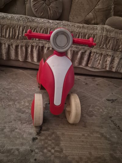 A red and white toy scooter with a silver circular accent on the handlebars is centered on a textured gray carpet. The scooter has two large, light-colored wheels visible. In the background, a plush, tufted sofa with ornate fabric and decorative pillows is slightly out of focus. The setting appears to be indoors, likely a residential room.