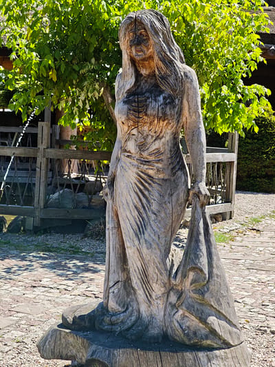 This image depicts a full-body wooden sculpture of a woman with long, flowing hair, dressed in a floor-length gown. The sculpture is situated outdoors on a cobblestone and gravel ground. In the background, there is a wooden fence with rope detailing and lush green foliage from trees and bushes, suggesting a garden or park setting. The sculpture itself is carved from a rough, weathered piece of wood, showing distinct grain patterns and natural imperfections. The woman is posed with one arm holding the folds of her dress. The lighting suggests a sunny day, casting shadows on the ground. The scene appears to be located in Horbów-Kolonia, Poland.