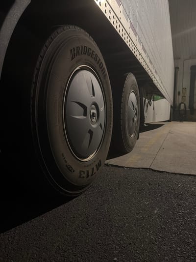The image shows the rear dual tires of a semi-trailer truck parked on an asphalt surface. The tires are branded "BRIDGESTONE M713" and have black hubcaps. The side of the trailer, which is white and metallic, is visible above the tires, featuring a repetitive pattern of small circular indentations. The lighting suggests it is nighttime or a dimly lit area. The location is identified as New Britain, United States of America, with the truck likely positioned in a loading dock or industrial area. No people are present in the frame, and the scene depicts a static, parked vehicle.