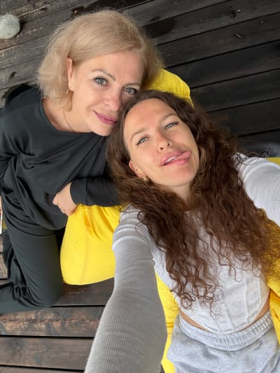 Two women are posing for a selfie on a wooden deck. The woman on the left has blonde hair and is wearing a black outfit. She is looking at the camera with a slight smile. The woman on the right has long, curly brown hair and is wearing a white top and grey sweatpants. She is sticking her tongue out at the camera. A bright yellow cushion is visible between them. The deck appears to be outdoors, possibly near a body of water, given the wooden planks and the overall casual setting. No specific landmarks or details indicate the location as Nizhnekayancha, Russia.
