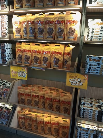 The image displays an indoor retail store environment, specifically a snack aisle or display, located in Tân Kỳ Commune, Vietnam. The scene is dominated by multiple tiers of shelving units and angled display racks filled with packaged food products.
The primary subject is a snack product labeled "Crispy Corn" with "Milk Chocolate" flavoring. These are packaged in light brown/tan stand-up pouches featuring red and white text, a clear window showcasing the chocolate-coated corn, and a carry handle cutout at the top. These pouches are densely stocked across three horizontal shelves and two lower angled display racks. A yellow shelf-edge sign beneath one shelf of "Crispy Corn" indicates the product name and a partial price, "36."
To the left of the "Crispy Corn" displays, on higher shelves, stacks of smaller, individually packaged dark and light-colored items, possibly cookies or biscuits, are visible. Below these, on an angled display, a yellow sign partially obscures packages, indicating "Pie" with cartoon cow graphics and "88."
To the right of the central display, vertically stacked packages labeled "Krispill" are arranged on shelves. These appear in both white and black packaging variations and seem to be cookie or wafer products. A prominent yellow sign advertising "Krispill" with an image of cookies and the price "28." is attached to a shelf.
The shelving units are constructed with dark vertical frames and light-colored wooden or laminate horizontal surfaces. Integrated horizontal LED strip lights illuminate the product displays from above each shelf. No individuals or active interactions are visible within the frame.