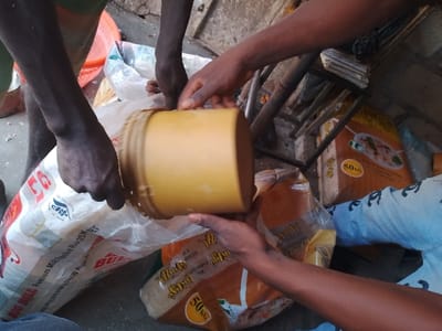 The image captures a close-up view of multiple dark-skinned hands involved in handling bulk rice, likely in a market or storage setting in Mgbuoshimini, Nigeria. At least three individuals' hands are visible, suggesting a collaborative activity.

In the foreground, a person on the left holds open a large white woven polypropylene bag. This bag is prominently branded with "BIG BULL" and "Premium Milled Parboiled Rice," along with "25KG NET" indicating its weight. Another pair of hands holds a light brown, cylindrical plastic bucket, which appears to be used for scooping or measuring the rice. The bucket is positioned over the opening of the "BIG BULL" bag, with some white rice grains visible around its rim, indicating the action of filling or emptying. A third set of hands is visible on the right, also seemingly assisting with the bucket or another bag.

In the background, more large bags of rice are stacked. One orange-brown bag is clearly visible, labeled "MARS RICE" and "50KG," suggesting different brands and sizes of rice are available. Another similar bag is partially obscured. Behind these bags, a simple metal structure or shelving unit is present, with what appears to be stacked papers or small items on top, implying a utilitarian space. A partial view of a red plastic basket is visible on the far left.

The lighting is bright and even, consistent with daytime conditions. The setting appears functional and practical, characteristic of a small commercial outlet or a food distribution point. No specific emotions are discernible from the hands, focusing the image on the task at hand. The floor appears to be concrete or a similar rough surface.