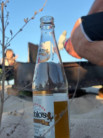 A close-up, eye-level shot shows a partially filled beer bottle in the foreground, sitting on sandy ground. The bottle is clear glass and has a white label with gold accents, featuring text that reads "HERMANOS Pablo's" and "LIGHT BEER" with an image of a lion. The beer inside is a golden amber color with a thin layer of foam near the top. In the blurred background, a person's legs are visible, wearing black athletic shorts with white trim and white socks. The person appears to be sitting on the sand. Behind the person, a dark, circular object, possibly a fire pit, is partially visible. The overall setting suggests an outdoor recreational area, perhaps a beach or park. The sky is clear and blue, indicating daylight. The location is identified as Povarovka, Russia.