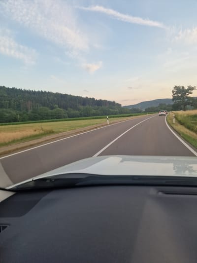 The image captures a view from inside a vehicle driving on a two-lane asphalt road in Jungingen, Germany. The road has clear white lane markings and is bordered by a grassy verge on the left and a field of dry grass and green crops on the right. In the distance, a dense forest covers a hillside, with a few scattered trees and some patches of exposed earth. The sky above is a pale blue with wispy white clouds, indicating either early morning or late afternoon. A white car is visible ahead on the right side of the road, and a red car is behind it. A large, mature tree stands prominently in the field on the right. The foreground shows the dashboard and windshield of the vehicle from which the photo was taken.