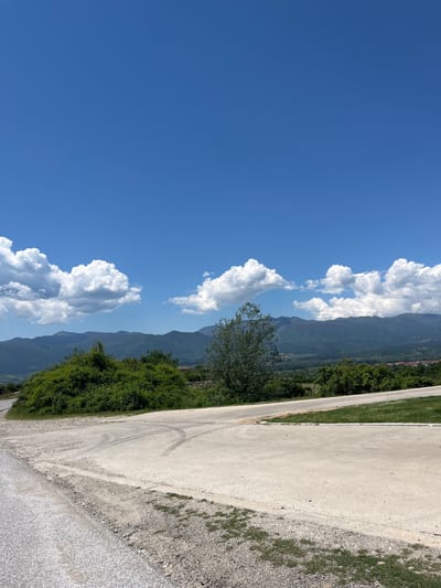 The image presents a landscape scene in Leshten, Bulgaria. A clear blue sky occupies the left portion of the view, punctuated by puffy white cumulus clouds. Mountains form the central vertical element, receding into the distance. Green vegetation covers the mountainside, with a solitary tree standing out. A narrow paved road stretches along the right edge of the frame, showing tire tracks. A dirt shoulder borders the road. No people are visible in the image.