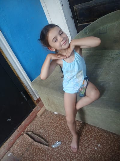 A young girl with brown hair is sitting on a couch with a blue wall behind her. She is wearing a light blue tank top with a "Frozen" theme and has her hands on her neck. A smartphone is resting on her right thigh. The floor has a speckled brown pattern, and there are some debris and a piece of torn linoleum. The environment appears to be indoors, possibly in a residential setting.