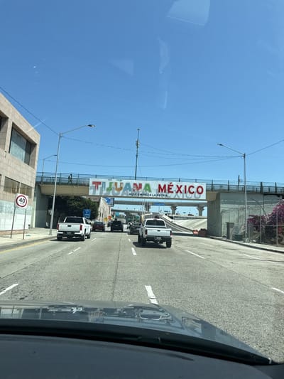 The image depicts a street in Tijuana, Mexico, specifically under a bridge that welcomes visitors to the city. The sign above reads "TIJUANA MEXICO" in large, colorful letters. Below this, in smaller text, it says "AQUI EMPIEZA LA PATRIA."

The view is from inside a vehicle, looking out through the windshield. In front of the vehicle, there are several other cars and trucks traveling in the same direction. The road is paved and marked with white lines.

To the left of the road, there is a building and a street sign indicating a speed limit of 40. To the right, there is a fence and some greenery.

The sky is clear and blue, suggesting it is daytime. The overall impression is of a busy street scene in a border city.
