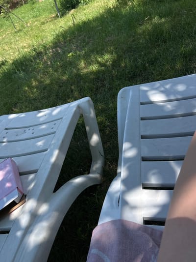 The image depicts a sun-drenched outdoor scene with two white plastic chaise lounges situated on a green grassy lawn. The lounges are positioned side-by-side, with the one on the left partially visible, showing its backrest and a pink box placed on it. The lounge on the right is more fully visible, with a pink towel draped over its seating area. A person's arm and leg are visible on the right side of the frame, indicating their presence on the right chaise lounge. The environment appears to be a garden or park, possibly in the city of Belo Pole, Bulgaria, given the presence of lush green grass and dappled sunlight filtering through tree branches, casting shadows on the lounges. The scene suggests a relaxed, leisurely atmosphere, likely during a warm day.