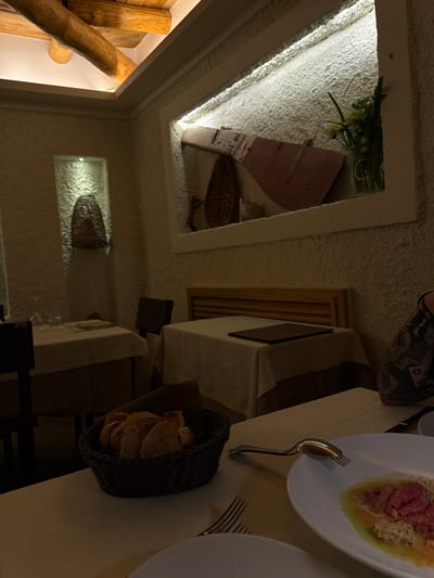 The image depicts a dining scene in what appears to be a restaurant in Agropoli, Italy. The perspective is somewhat skewed, likely from a handheld shot.

Foreground: A table setting is visible, covered with a beige tablecloth. A white plate holds food, possibly a tartare or a similar dish with meat and other ingredients. A fork and spoon are placed near the plate. Next to the plate is a basket with bread.

Background: The restaurant's interior design is visible. The walls and ceiling have a textured, possibly stucco, finish. There are decorative features set into the walls, including what looks like a display with a paddle and plant. There is an opening revealing some wooden beams. Two tables with white linens are placed near each other. There's also a glimpse of a person's clothing at the upper part of the image.