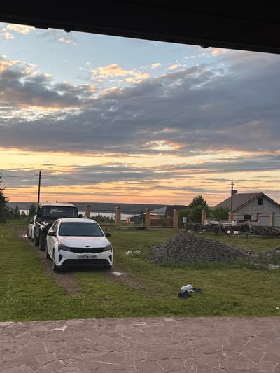 The image depicts a rural or semi-rural setting, likely in or near the village of Пепеляево, Russia, during a sunset. In the foreground, a paved patio area made of irregular, reddish-brown stones is visible. Behind this, a grassy yard contains a dirt path leading to a line of parked vehicles. The closest vehicle is a white Kia sedan with a license plate reading "KO07KE 159". Behind it are at least two dark-colored SUVs or off-road vehicles.
The midground shows a residential construction area. A large pile of dark gravel or crushed stone is prominent. Further back, a fence constructed of brick pillars and wire mesh separates the property from a building under construction, appearing to be made of concrete blocks with a pitched roof. Trees and other foliage are scattered throughout the landscape.
The background features a distant body of water, likely a lake or river, stretching across the horizon. The sky is dominated by a dramatic sunset with a mixture of scattered white and grey clouds illuminated by warm orange and yellow light. Power lines and a utility pole are visible against the sky, indicating the presence of infrastructure. No people are visible in the scene.