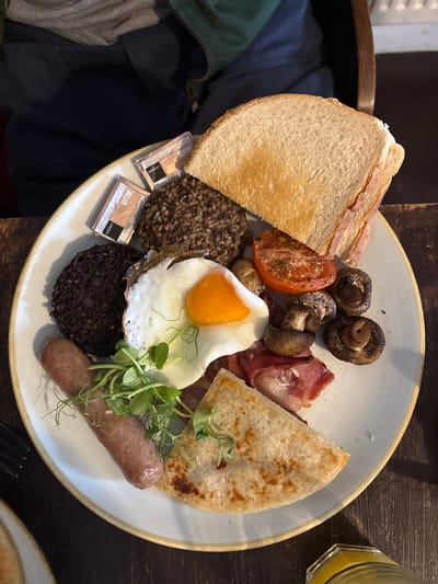 A full breakfast plate is presented on a rustic wooden table next to a half-full glass of orange juice. The plate contains toast slices, mushrooms, grilled tomato, bacon, a fried egg, a potato scone, some green garnish, a sausage, and black pudding, served with two packets of butter. A chair and folded clothes are visible next to the plate. The location is assumed to be Carbost, United Kingdom.