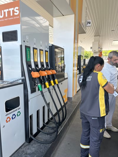 The scene depicts a gas station, likely in Gaziosmanpaşa, Turkey, based on the Turkish text and the style of the gas pump. A woman wearing a uniform is interacting with a customer near a gas pump. The pump has a display showing numbers 5 through 8, and nozzles indicating different fuel types. A recycling receptacle is visible on the pump. The station has a covered roof, and daylight suggests an outdoor setting.