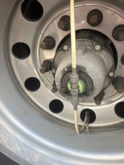 The image shows a close-up view of a truck wheel hub, likely a commercial vehicle. The wheel is made of silver metal and has several circular cutouts. The central hub assembly is also made of metal and has various markings, including "HUB-SEAL" and "STEMCO". There is a green colored cap or seal visible in the center of the hub. A light-colored tube, possibly a tire pressure monitoring system line, extends from the hub towards the viewer. The wheel appears to be attached to a vehicle, and the background suggests an outdoor setting. There are no people or discernible actions taking place in the image. The specific location within the city of Lebanon, United States of America cannot be determined from the image alone.