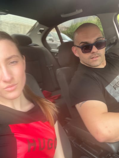 Two individuals, a woman and a man, are seated in the interior of a car, appearing to be in motion. The woman, positioned in the foreground on the left, has light skin, long light brown hair pulled back, and is wearing a black t-shirt with a prominent red section displaying the "HUGO" logo in dark lettering. The man, in the right foreground, has a bald head, is wearing dark sunglasses, and a black t-shirt with a grey graphic that includes the word "PERF". His right arm, showing some red marks on the elbow, rests on the car's center console. The background reveals the car's interior, including dark seats, and through the windows, glimpses of outdoor scenery with buildings and vegetation, suggesting an urban or suburban environment. The scene is captured in what appears to be daylight. The location is specified as Vlahovo, Bulgaria.