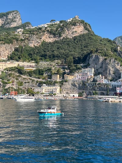 The image shows a view from the water of the coastal town of Pogerola, Italy. In the foreground, a small turquoise and white boat is on the water. In the middle ground, several larger white boats and many smaller boats are docked in a harbor. Behind the harbor, buildings cascade down the steep, rocky cliffs. The buildings are various pastel colors, including yellow, pink, and white, and have red-tiled roofs. Green trees and vegetation cover the lower slopes of the cliffs, and the upper slopes are bare rock. The sky is clear and blue. The overall impression is of a picturesque Italian coastal town.