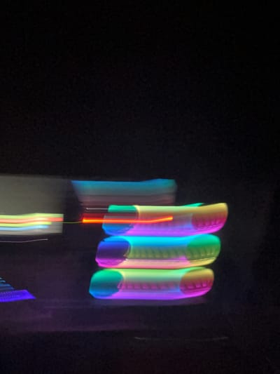 The image is a long exposure photograph of several glowing, cylindrical objects stacked vertically. The objects are illuminated with a spectrum of colors, including blues, greens, yellows, oranges, and purples. The lighting creates streaks and blurs due to motion during the exposure. The background is predominantly black, suggesting a dark or nighttime environment. On the left side of the image, there are faint, horizontal streaks of light in various colors, possibly indicating other moving lights or objects out of focus. The overall impression is abstract and artistic, with a focus on light and color trails. This photograph was taken in Stallings, North Carolina, United States of America.
