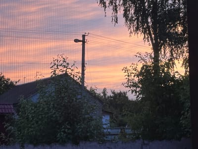 This media file captures a serene sunset scene in Seredivka, Ukraine. The sky is painted with soft hues of orange and pink, streaked with thin clouds that create a textured, layered effect. A power pole with a street lamp stands tall against the colorful sky, its wires crisscrossing the frame.
In the foreground, dark green foliage from trees and bushes partially obscures a weathered, light-colored house with a dark roof. The house appears to be a rural dwelling, likely residential. A white picket fence is visible behind the bushes, hinting at a yard or garden area. The overall impression is one of quiet domesticity set against the dramatic backdrop of the setting sun.
The time of day is clearly dusk, with the sun low on the horizon and casting a warm glow. The weather appears calm, with no strong winds suggested by the foliage. The presence of the power pole and the house with a tiled roof indicate a settled, possibly rural or suburban, location. There are no people visible in the image, and no text is present. The scene evokes a sense of peace and tranquility, characteristic of a quiet evening in the countryside.
