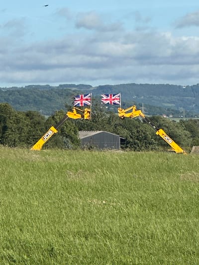 In a rural landscape, two yellow JCB construction vehicles are positioned, each hoisting a Union Jack flag. The vehicles are angled, with their booms extended upwards and outwards. The foreground is dominated by a field of tall, green grass. In the mid-ground, behind the grass, are trees and a dark, rectangular building, possibly a barn or agricultural shed. Beyond this, a hilly, tree-covered landscape stretches into the distance under a partly cloudy sky. A single bird is visible in flight in the upper left quadrant of the sky. The overall impression is of a festive or celebratory display set in the countryside near Tewkesbury, United Kingdom.