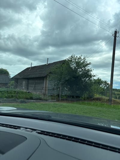 The image shows a cloudy, overcast sky above a rural scene. In the foreground, the dashboard of a car is visible, suggesting the photo was taken from inside a vehicle. The scene outside the car includes a weathered wooden house with a dark, shingled roof and a small window. A green, bushy tree grows to the right of the house, partially obscuring it. Further in the background, there are more trees and vegetation. A wooden utility pole with wires stretches upwards on the right side of the image. The ground is covered in grass, and some fencing is visible. The overall impression is of a simple, rural dwelling in a natural setting. The location is identified as Kryŭkaŭski sel’ski Savet, Belarus.